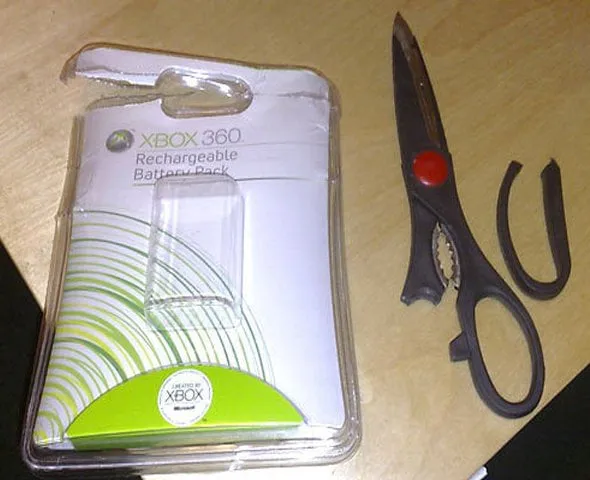 Xbox 360 rechargeable battery pack still sealed in clamshell plastic, next to kitchen scissors with a snapped-off handle.